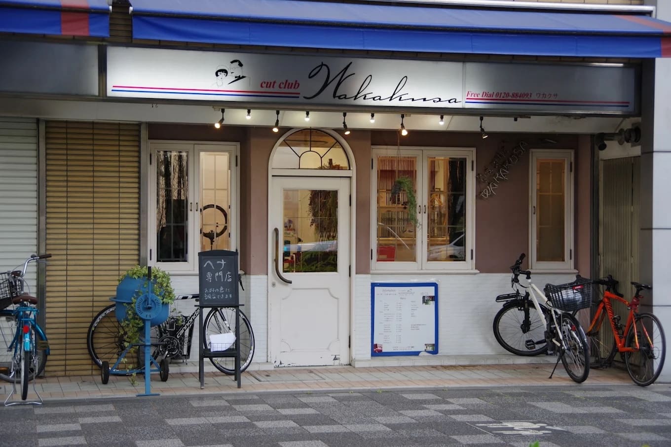 这家商店的白色双门上有一个写着“cut club Wakana”的牌子，并提供捷径和指甲花服务。 夹在窗户和自行车停车场之间，入口附近有蓝色的自行车架、室内植物以及菜单和标志。