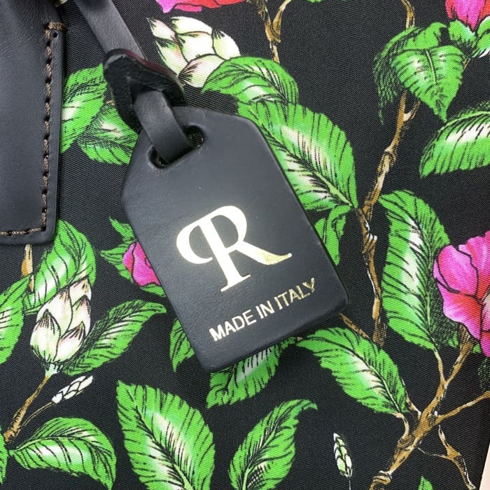 带有金色“R”和意大利文字的黑色 ROBERTA PIERI 行李牌悬挂在带有绿叶和粉红色花朵的花卉面料上，与迷你行李袋相得益彰，具有永恒的意大利优雅。